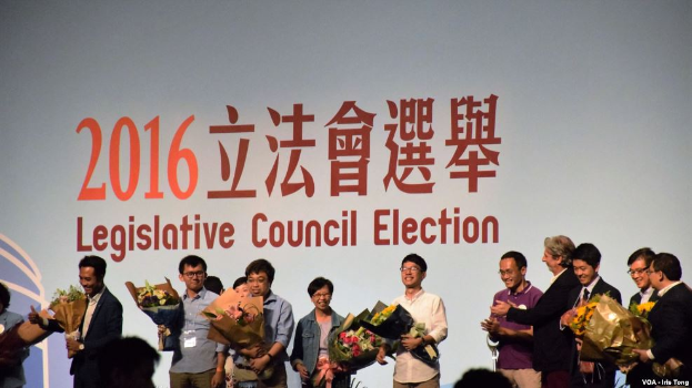 Eine Gruppe von Menschen auf einer Bühne mit Blumensträußen, umgeben von einer Zuschauermenge und einem Bildschirm mit Text und Logo für die Wahl des Legislativrats 2016 im Hintergrund.