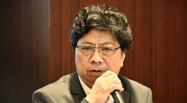 Ein Mann im Anzug mit Mikrofon zu sehen, der scheinbar in einer hitzigen Diskussion mit einer hölzernen Wand im Hintergrund ist.