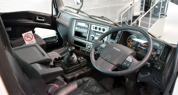Innenansicht eines Truck-Cockpits mit Lenkrad, Armaturenbrett, Sitzen, Ganghebel, Tacho, Scheibenwischer, Seitenspiegel und weiteren Komponenten sowie Geländern und zusätzlichen Fahrzeugen im Hintergrund.