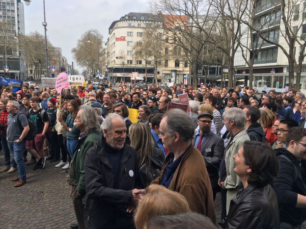 Eine große Gruppe von Menschen vor einer Menge mit Schildern, mit Fahrzeugen, Bäumen, Laternenmasten und Gebäuden im Hintergrund, wahrscheinlich während einer Demonstration in Berlin, Deutschland.