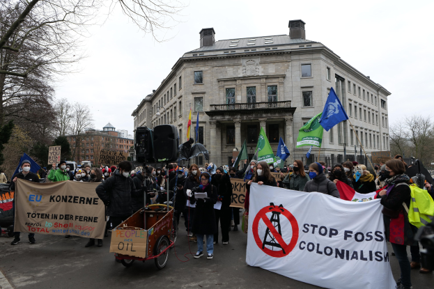 Große Gruppe von Menschen marschiert mit Transparenten und Fahnen durch die Straße, mit einem Fahrzeug im Vordergrund, protestiert gegen fossile Brennstoffe; Bäume, Gebäude mit Fenstern und ein klarer blauer Himmel im Hintergrund.