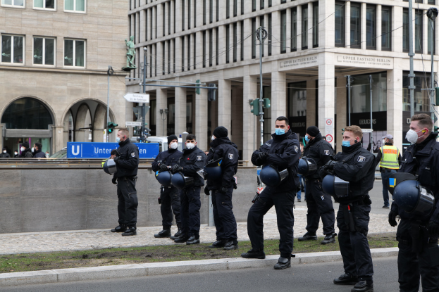 Eine Gruppe von Polizisten in schwarzen Uniformen und Masken steht vor einem Berliner Gebäude mit Glasfenstern und Säulen, einige halten Helme, mit Laternenmasten, Verkehrsampeln, Texttafeln, einer Statue und anderen Menschen im Hintergrund.
