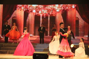 Eine Gruppe von Menschen auf einer Bühne mit Stufen und einem Hintergrundbild, beleuchtet von Scheinwerfern und mit einem Lautsprecher unten, was eine Tanzvorstellung suggeriert.