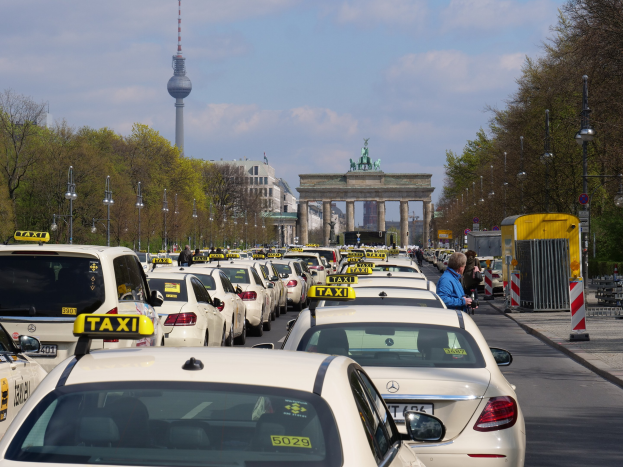 Eine belebte Straße in Berlin mit zahlreichen geparkten Taxis, Fußgängern auf dem Gehweg, Laternen, Bäumen, Gebäuden und einem fernen Bogen mit Statuen und Turm unter einem bewölkten Himmel.