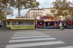 Eine belebte Stadtstraße mit Zebrastreifen und Karussell, mit Menschen, Ständen, Bäumen, Gebäuden, Bannern, Laternen und einer Ampel unter einem bewölkten Himmel.