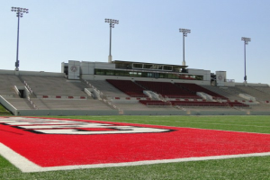 Fußballfeld mit rot-weißem Streifen auf saftig grünem Rasen, umgeben von Tribünen, Treppen, Geländern, Pfosten, Lampen und einem Gebäude unter einem klaren blauen Himmel.