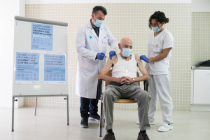 Ein älterer Mann sitzt in einem Krankenhauszimmer auf einem Stuhl, während ihm zwei maskierte und handschuhgetragene Ärzte ein Vakzin verabreichen, mit einer Tafel links und Gegenständen auf einer Plattform rechts.