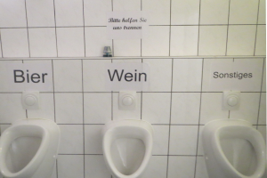 Drei weiße Toiletten an einer Wand montiert mit Schildern darüber, die 'Bier Wein' und 'Sonstiges' lesen.