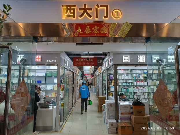 Innenseite einer chinesischen Apotheke mit Glaswänden, Regalen voller Artikel, verstreuten Kartons, einer Pflanze links und einem Namensschild oben.