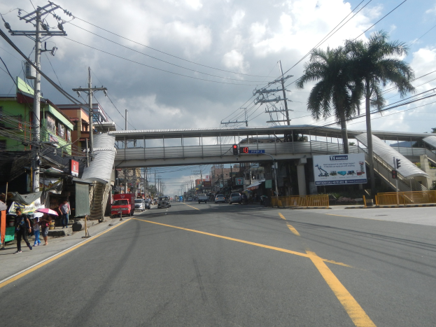 Stadtstraße mit Fahrzeugen, Fußgängern, Strommasten, Gebäuden, Bäumen, einer Brücke, einer Ampel und einem bewölkten Himmel.