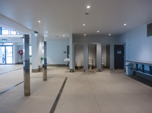 Große Öffentliche Toilette mit mehreren Toiletten, tragenden Säulen, einem Mülleimer, einer Tür, einem Schild, einem angebrachten Feuerlöscher und Deckenleuchten auf dem Dach.