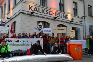 Eine Gruppe von Menschen mit Schildern und Plakaten steht vor einem Gebäude, mit zwei Personen im Vordergrund und einem Müllcontainer rechts daneben, während einer Demonstration in Deutschland, mit Gebäuden, Fenstern, Lichtern und Schildern im Hintergrund.