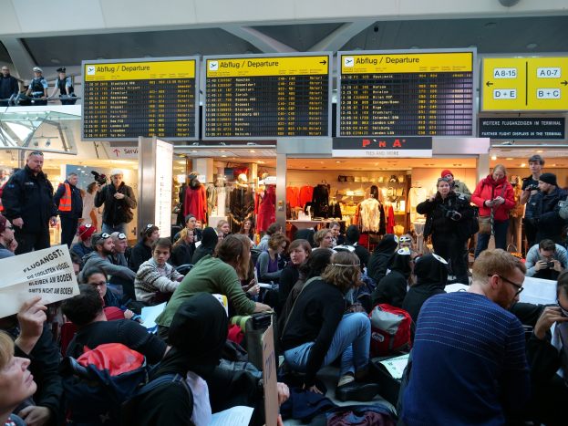 Eine große Gruppe von Menschen in einem Flughafen, einige mit Taschen und Papieren sitzend, andere stehend, mit Schildern, Puppen in Kleidern und Deckenlampen im Hintergrund, was auf eine Demonstration hinweist.