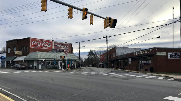 Stadtstraße mit Ampel an einer Kreuzung, Fahrzeuge, Gebäude mit Namenschildern, Strommasten mit Drähten, Bäume und ein bewölkter Himmel im Hintergrund.