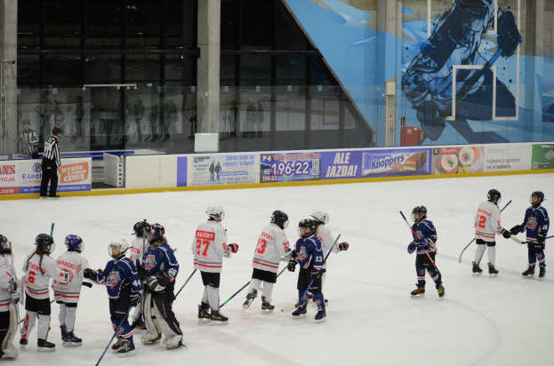 Group of young people playing ice hockey on an indoor rink, wearing helmets and holding sticks, with a person standing on the left side and banners, pillars, and glass walls in the background.