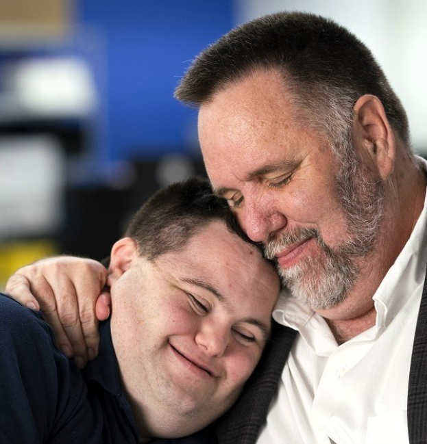 Zwei Personen, ein Mann in einem weißen Hemd und schwarzer Blazer und ein Junge, umarmen sich in einer herzlichen Umarmung vor einem unscharfen Hintergrund.
