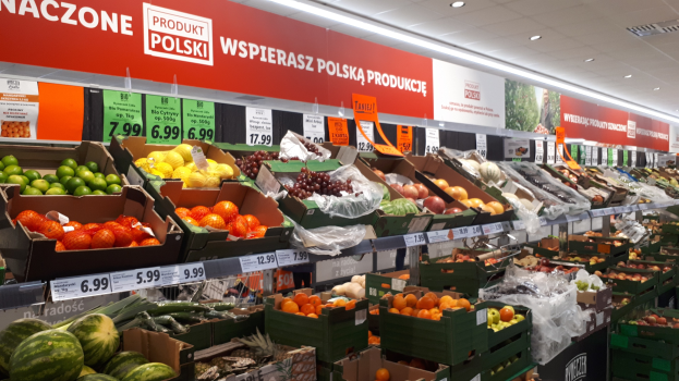 Ein Geschäft mit verschiedenen Obst- und Gemüsesorten in Kartonschachteln, jeweils mit Preisschildern, beleuchtet von Deckenlampen und mit Informationsschildern an den Wänden.