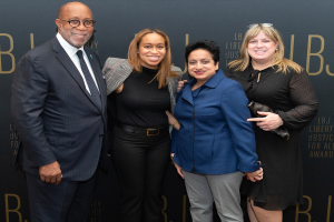 Eine Gruppe von vier Personen, die auf einem roten Teppich stehen, lächeln und für ein Foto posieren, mit einem 'Liberty Justice for All Awards'-Schild im Hintergrund.