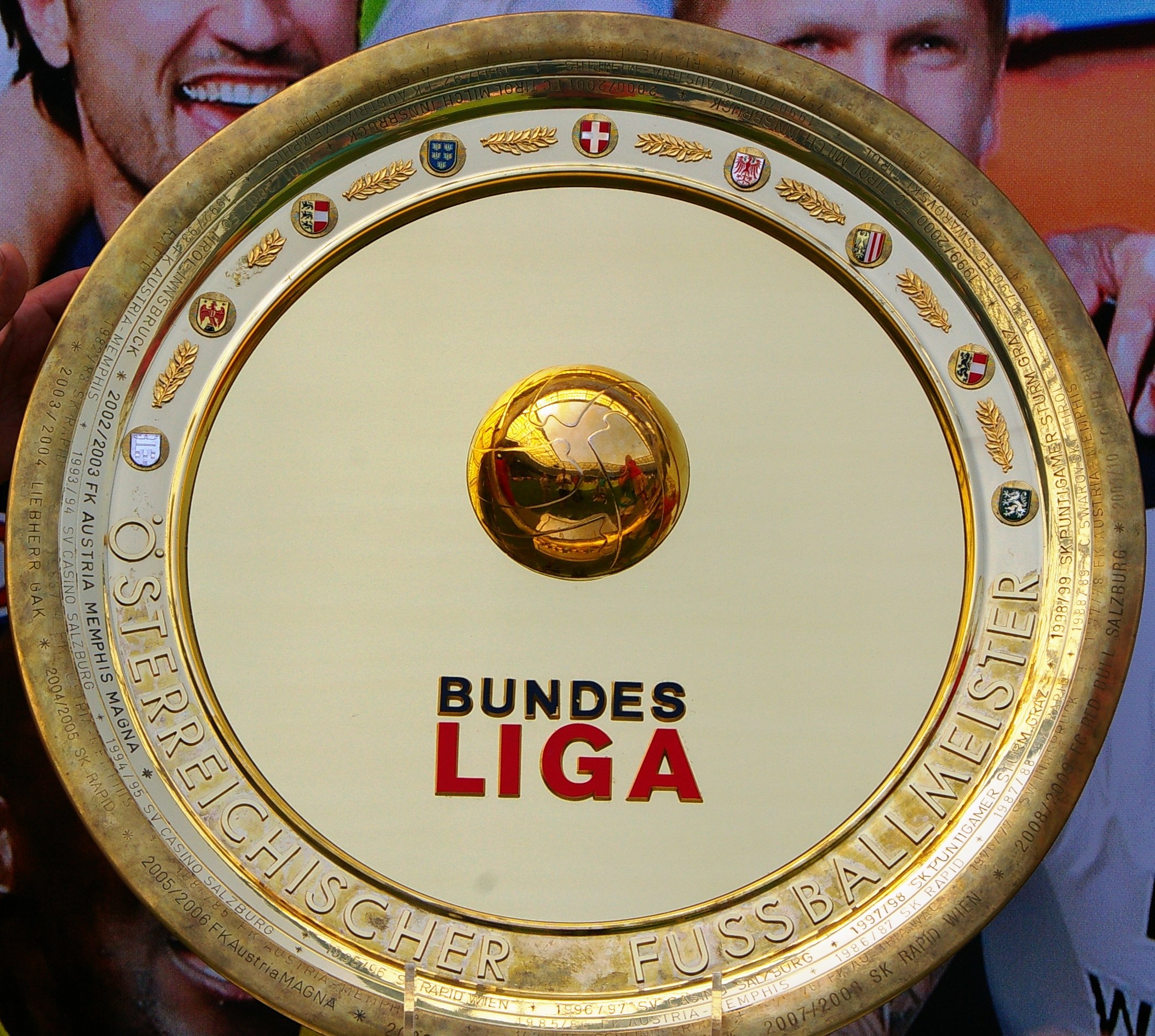 Goldener Schild mit "Bundesliga"-Text in der Hand eines Mannes, Hintergrundbanner mit drei Personenbildern.