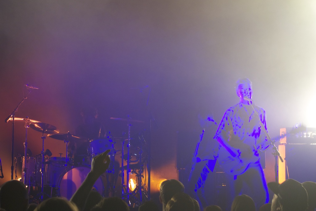Ein Mann spielt Gitarre vor einem Mikrofon, mit Musikinstrumenten und Lichtern im Hintergrund.