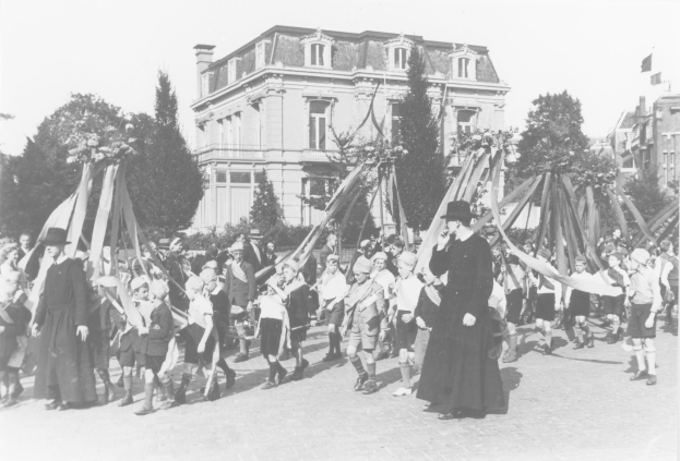Schwarz-weißes Bild einer Gruppe von Menschen, die eine Straße entlanggehen und Fahnen schwenken, mit Hüten, Bäumen, Gebäuden und einem Maibaum im Hintergrund.