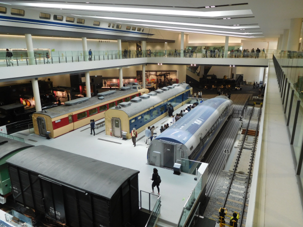 Eine große Museumsausstellung zeigt verschiedene Züge in unterschiedlichen Größen und Farben, mit Menschen, die sie von Boden, Geländern und Säulen unter Deckenbeleuchtung betrachten.