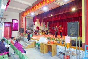 Eine Bühne mit zwei singenden Personen, eine Band im Hintergrund, rote Vorhänge, eine Lampe, ein Lautsprecher rechts und ein Publikum auf Stühlen sitzend.