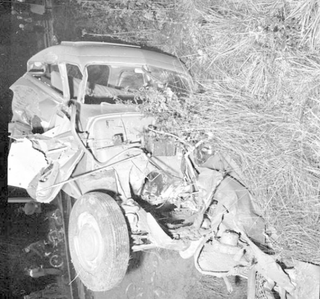 Schwarz-weiß-Foto eines schwer beschädigten Autos mit eingedrückter Front und zersplitterten Fenstern am Rande einer Straße, mit einem Baum und verstreuten Gegenständen im Hintergrund.