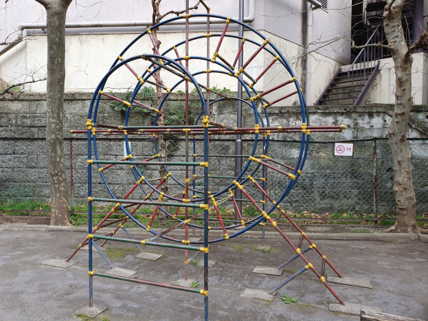 Ein Spielplatz mit einer große Metallkonstruktion, umgeben von Bäumen und einer Maschendrahtzaun-Mauer, mit Treppen, die zu einem Gebäude mit Fenstern und Rohren führen, einem Schild an einem Pfahl und grasbewachsenem Boden.