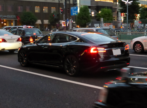 Schwarzer Tesla Model S fährt auf einer nächtlichen Stadtstraße mit anderen Fahrzeugen, Gebäuden, Bäumen und Fußgängern im Hintergrund.