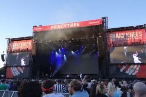 Eine große Menschenmenge steht vor einer hell erleuchteten Bühne beim Shaky Knees Music Festival, auf der Musiker hinter einem Geländer unter einem klaren blauen Himmel spielen.