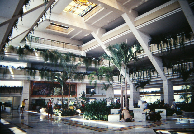 Ein geräumiger Hotellobby-Atrium mit Menschen, Grünpflanzen, Säulen und Beleuchtung unter einer beleuchteten Decke.