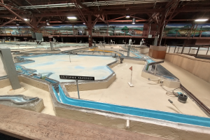 Ein großer Indoor-Wasserpark mit zahlreichen Wasserrutschen, Pfählen, Schildern, Deckenleuchten und Wandgemälden, der wie ein Minigolfplatz gestaltet ist.