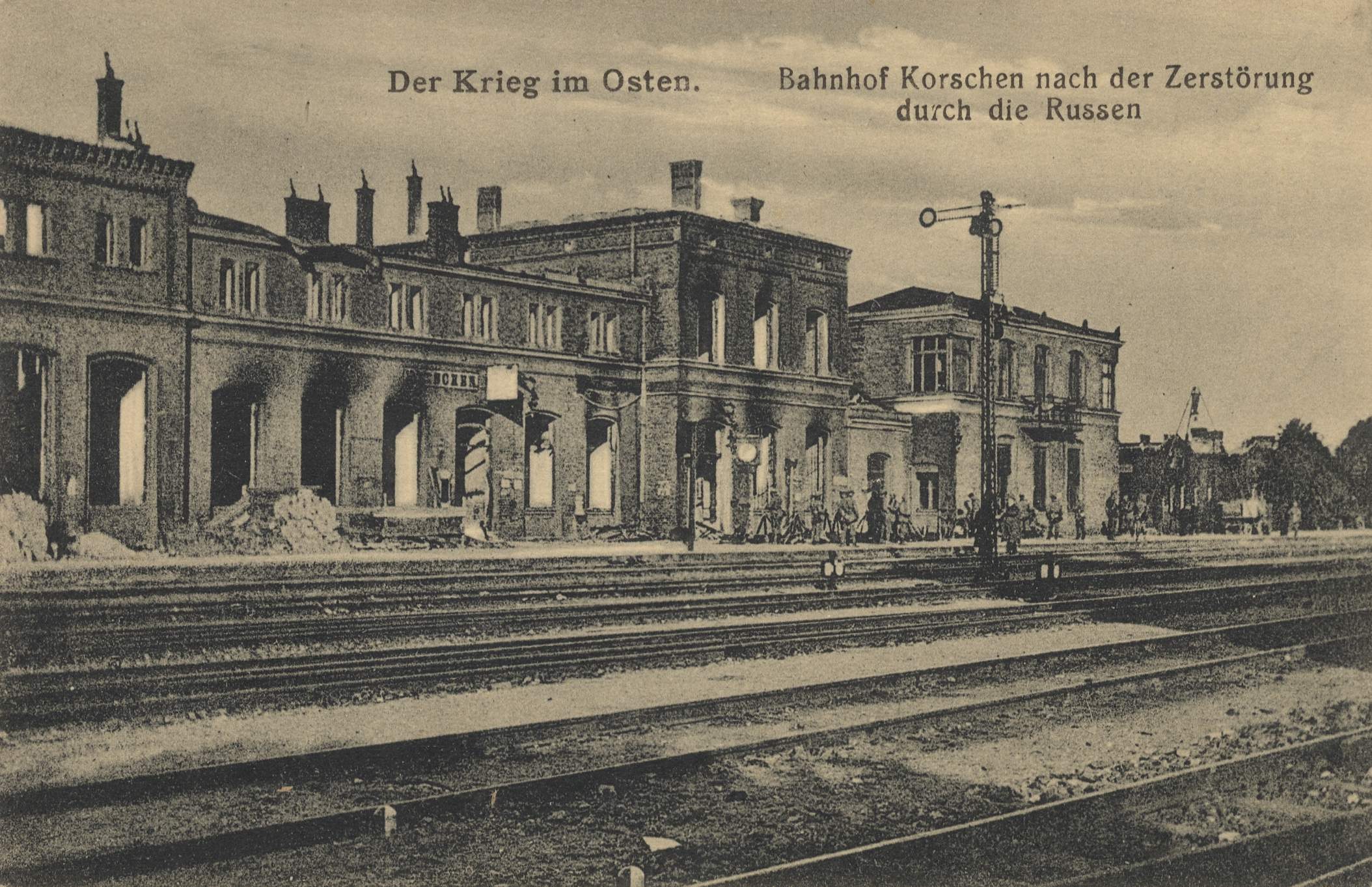 Schwarzes und weißes Foto des Bahnhofs Korschen in Ostern, Deutschland, das Schienenstränge, Bahnhofsgebäude, Menschen, Bäume und Himmel zeigt.
