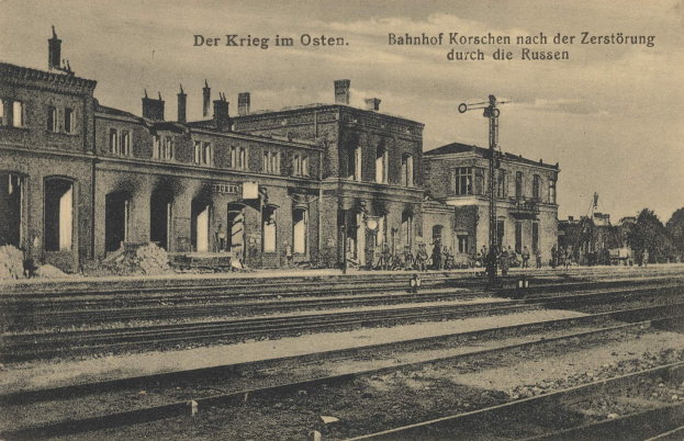 Schwarzes und weißes Foto des Bahnhofs Korschen in Ostern, Deutschland, das Schienenstränge, Bahnhofsgebäude, Menschen, Bäume und Himmel zeigt.