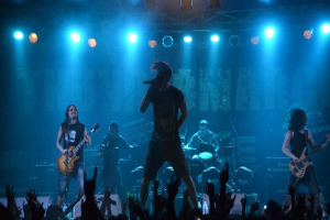 Eine Gruppe von Musikern, die auf einer hell erleuchteten Bühne in der O2 Arena in London spielen, darunter einige mit Gitarren, einer mit Mikrofon und einer, die im Hintergrund Schlagzeug spielt, während unten Hände des Publikums zu sehen sind.