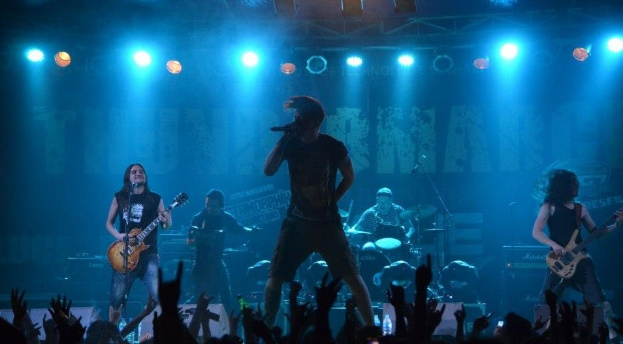 Eine Gruppe von Musikern, die auf einer hell erleuchteten Bühne in der O2 Arena in London spielen, darunter einige mit Gitarren, einer mit Mikrofon und einer, die im Hintergrund Schlagzeug spielt, während unten Hände des Publikums zu sehen sind.