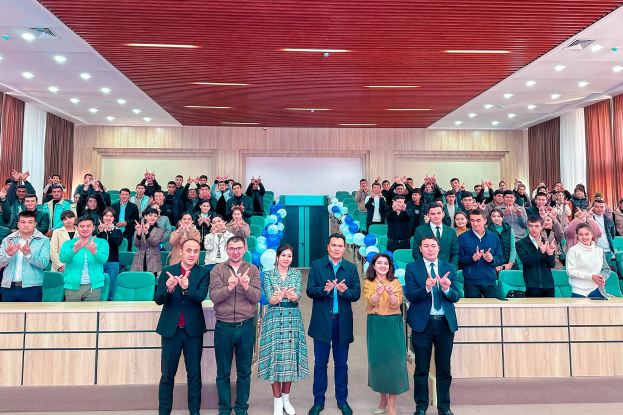 Eine Gruppe von Menschen steht vor einer Menge, einige klatschen, in einem Raum mit Stühlen, Tischen, Ballons, Vorhängen und Deckenleuchten, feiernd die Eröffnung einer neuen Schule.