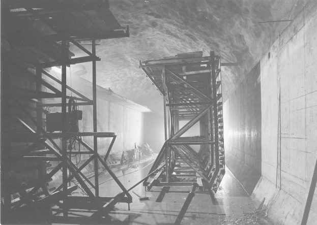 Schwarzes und weißes Foto eines Tunnels mit einer zentralen Metallstruktur, Metallstangen auf der linken Seite, einer Wand auf der rechten Seite und Text unten.