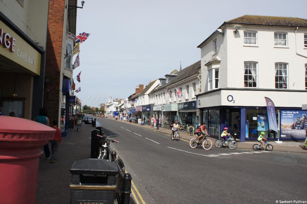 Eine Straße mit Gebäuden auf beiden Seiten, die Radfahrer auf der Straße, Fußgänger und Fahrzeuge auf einem linken Gehweg, Flaggen auf den Gebäuden und ein Banner vor einem Geschäft auf der rechten Seite zeigt.