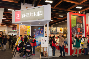 Eine große Gruppe von Menschen auf einer Messe vor einem Stand mit Stühlen, Schildern mit der Aufschrift "Book Republic Hong Kong Book Fair", Banner mit Text und Bildern und einer Decke mit Deckenleuchten.