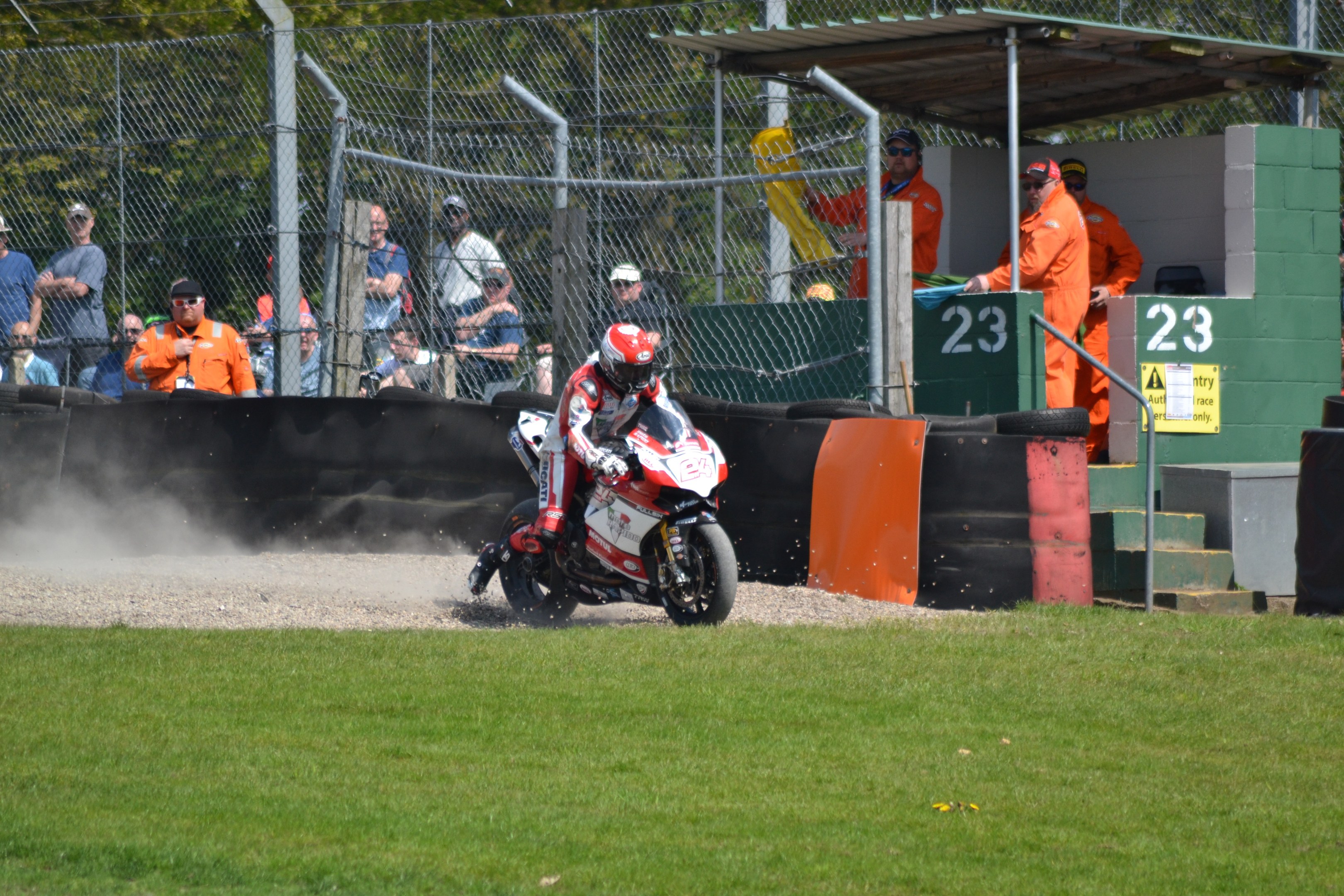 Ein Motorradfahrer in Helm und Handschuhen fährt über ein grasbewachsenes Feld mit Zuschauern, Absperrungen, Treppen und einem nummerierten Schuppen im Hintergrund.