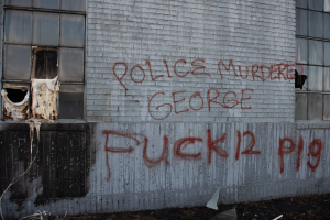 Graffiti an einer Gebäudewand mit der Aufschrift "Polizei ermordet George Fuck 12 P19" mit Fenstern und verstreuten Gegenständen davor.