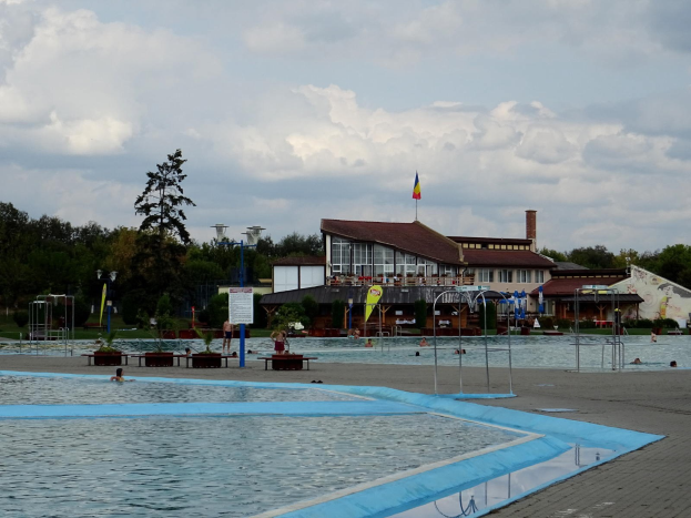 Ein großes Schwimmbecken mit schwimmenden Menschen, umgeben von Pfählen, Bänken, Topfpflanzen, einem Schild, einem Fahnenmast mit einer Flagge, einem Gebäude mit Fenstern, Straßenlaternen, einer Baumgruppe, unter einem bewölkten Himmel.