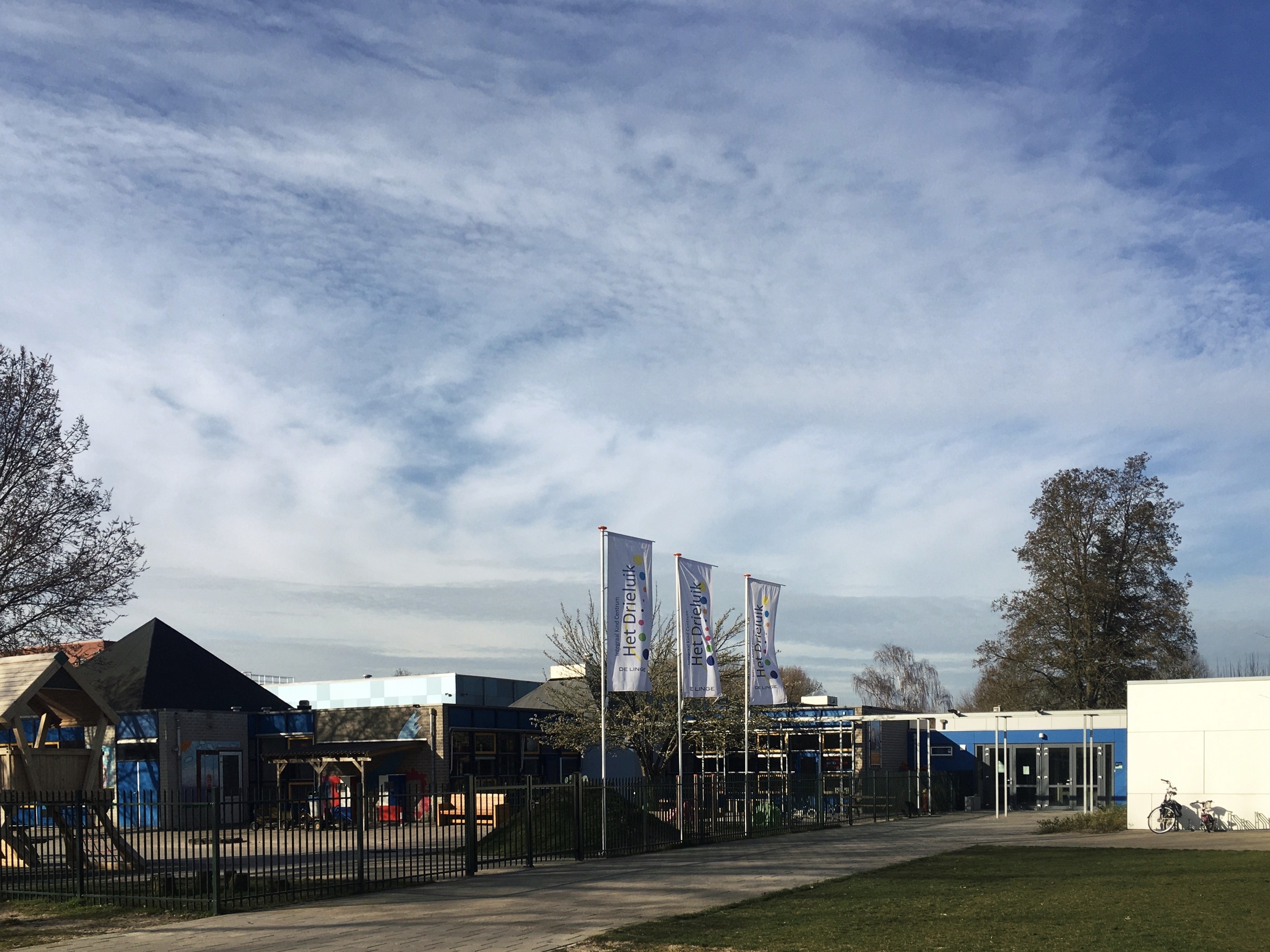 Großes Gebäude mit Metallzaun, Rasenfläche, Weg, Bäumen, Bannern, Fahrrad, nahen Häusern und bewölktem Himmel, identifiziert als neuer Spielbereich für Kinder in der Schule.