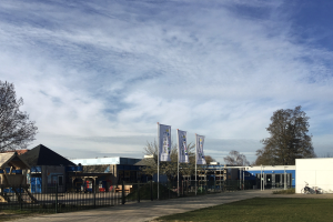Großes Gebäude mit Metallzaun, Rasenfläche, Weg, Bäumen, Bannern, Fahrrad, nahen Häusern und bewölktem Himmel, identifiziert als neuer Spielbereich für Kinder in der Schule.