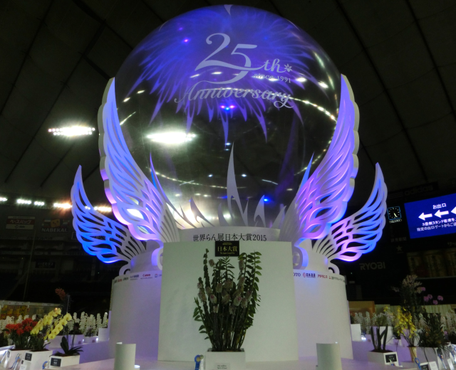 Ein großer Kristallball mit Flügeln oben drauf, umgeben von Blumenbouquets, Namensschildern und anderen Gegenständen auf dem Boden, mit Tafeln, Lichtern und einem Bildschirm im Hintergrund bei einer 25-jährigen Jubiläumsfeier.