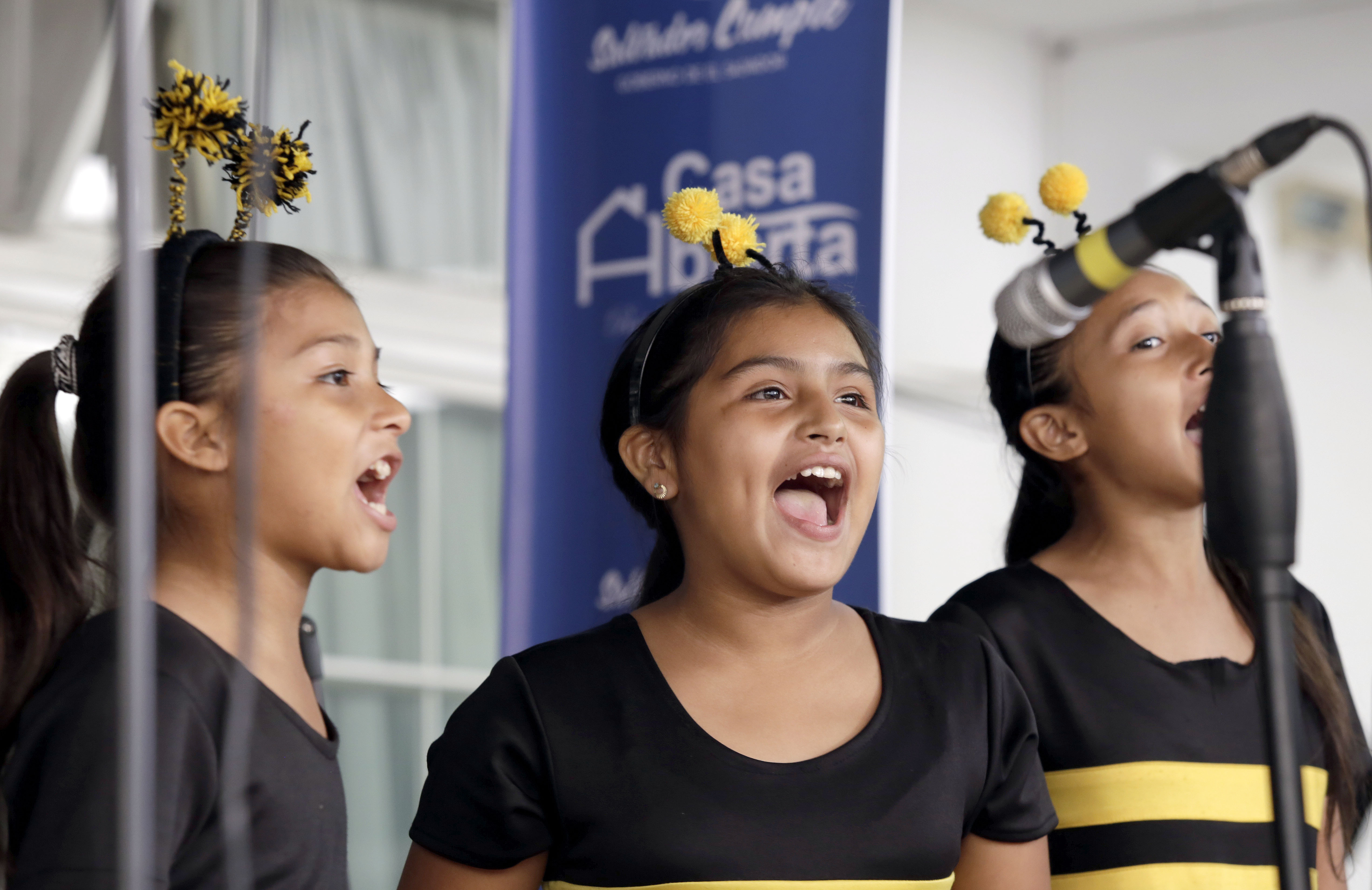 Drei junge Mädchen in schwarzen und gelben Kleidern, die in ein Mikrofon singen, mit einem blauen Banner und einer weißen Wand im Hintergrund.