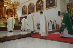Eine Gruppe von Priestern geht über einen roten Teppich in einer Kirche, mit Menschen, die auf dem Boden vor ihnen und auf der rechten Seite stehen, und Fotorahmen an der Wand im Hintergrund.