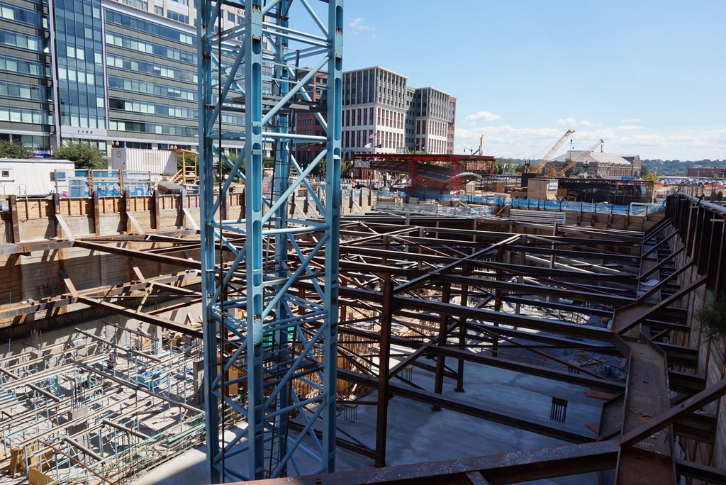 Das Bild zeigt eine Baustelle mit Gebäuden und Büros links, Eisenstangen und Maschinen rechts und diverser Ausrüstung und Kränen in der Mitte, unter einem Himmel oben rechts.